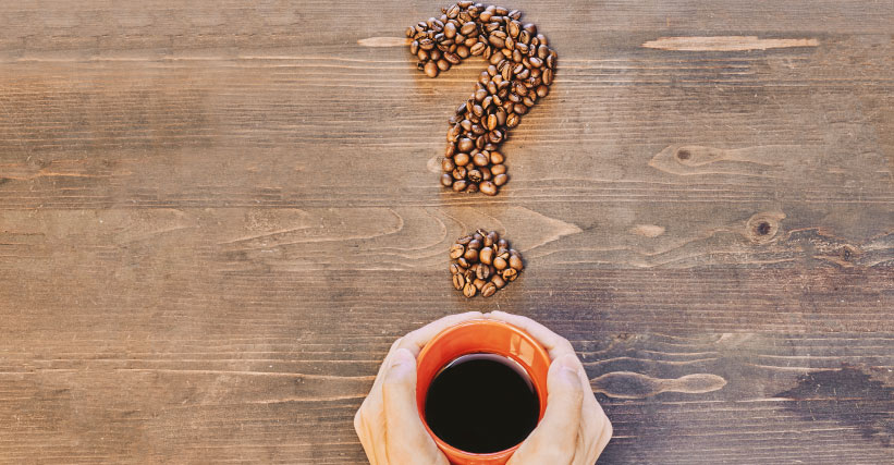 Mitos e verdades sobre café - Pessoa segurando xícara de café sobre mesa de madeira com ponto de interrogação formado por grãos de café.