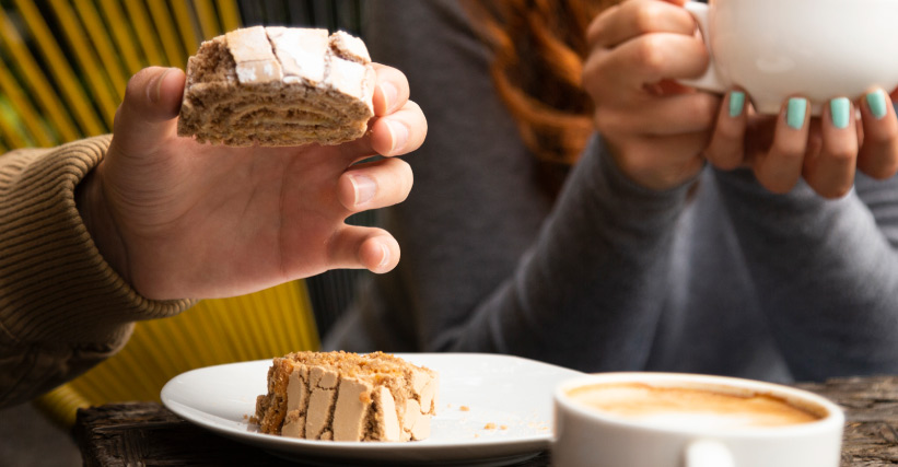 O que combina com café - Duas pessoas tomando café e comendo doce em ambiente descontraído, com foco nas mãos segurando xícara e fatia de bolo.