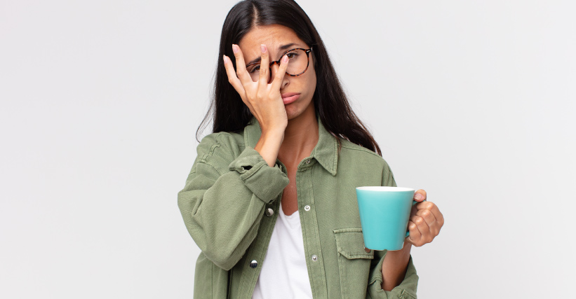 Café Fake = Mulher com expressão cansada cobrindo o rosto parcialmente com a mão enquanto segura uma caneca azul, sugerindo frustração.