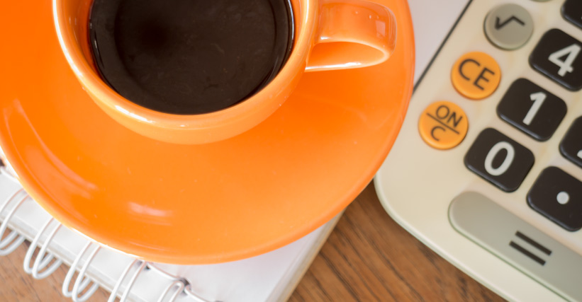 Porque o café está caro - Xícara de café preto sobre pires laranja, ao lado de uma calculadora e um caderno espiral, simbolizando análise de custos.