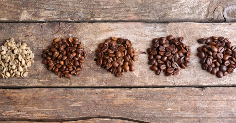 Qual a melhor torra de cafe - Cinco porções de grãos de café exibindo diferentes níveis de torra, organizadas em uma superfície de madeira rústica.