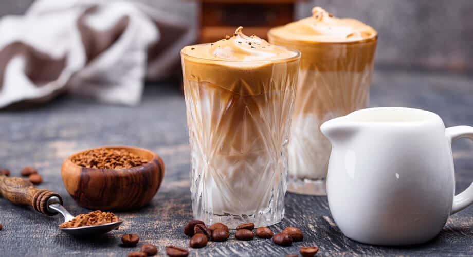 café com leite condensado servido em um copo de vidro, posto em uma mesa cinza ao lado de grãos de café