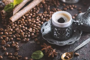 xícara de café sobre uma bandeja metálica, rodeada de grãos de café, paus de canela, folhas de hortelã e estrelas de anis