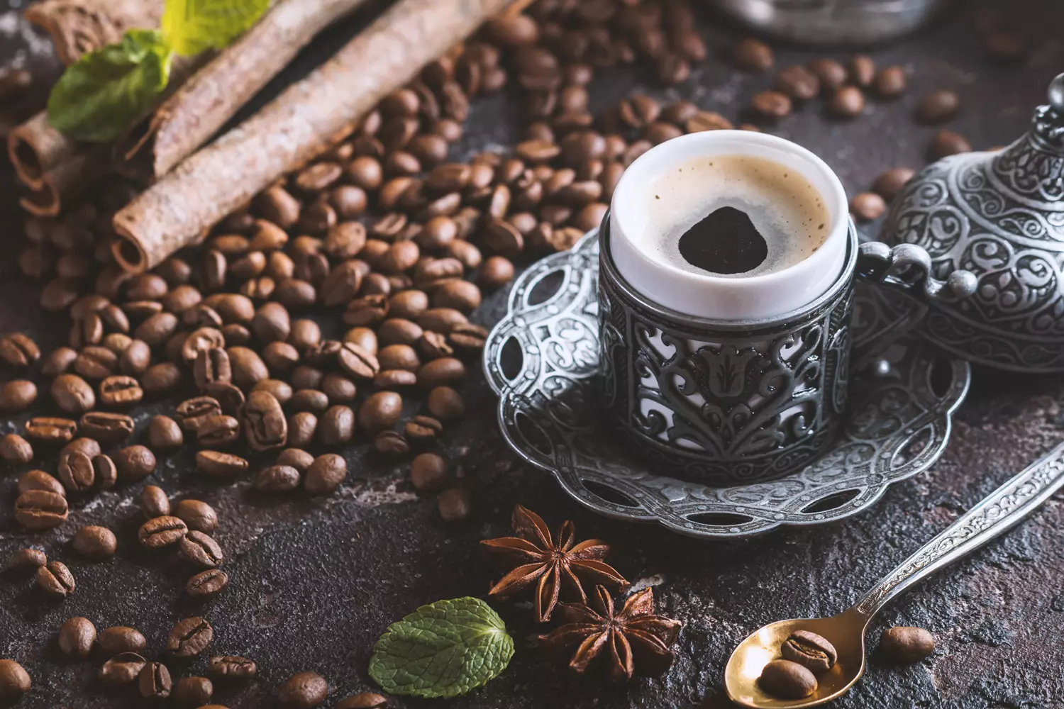 xícara de café sobre uma bandeja metálica, rodeada de grãos de café, paus de canela, folhas de hortelã e estrelas de anis